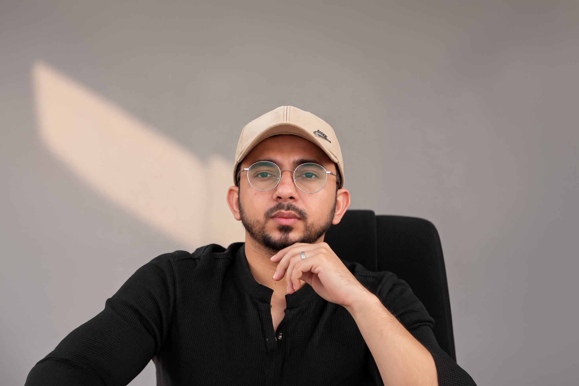 Muhammad Kashif in a beige cap, round glasses and a black shirt, resting his chin on his hand
